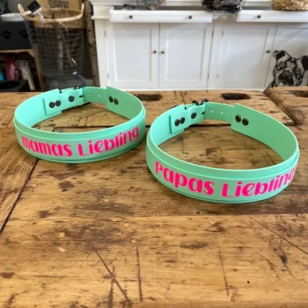 Zwei Hundehalsbänder aus dem Material Biothane. Auf den Halsbändern ist eine Aufschrift. Auf dem einen Halsband steht Mamas Liebling und auf dem anderen Halsband steht Papas Liebling in pink. Die Halsbänder sind mint Farben.